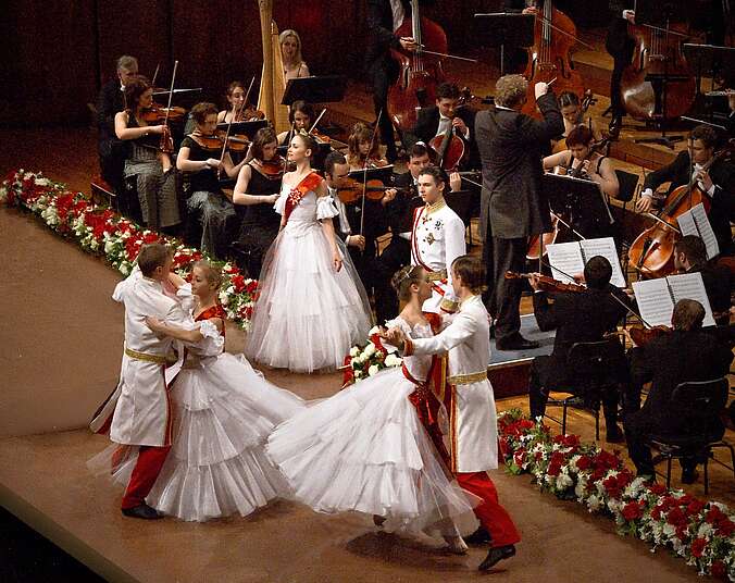 Ein Orchester spielt ein Stück während zwei Paare in weißem Kleid und weißem Anzug vor ihm einen Walzer tanzen und und sich drehen. Ein weiteres Paar steht vor Ihnen und blickt ins Publikum. Sie tragen das gleiche Kostüm.