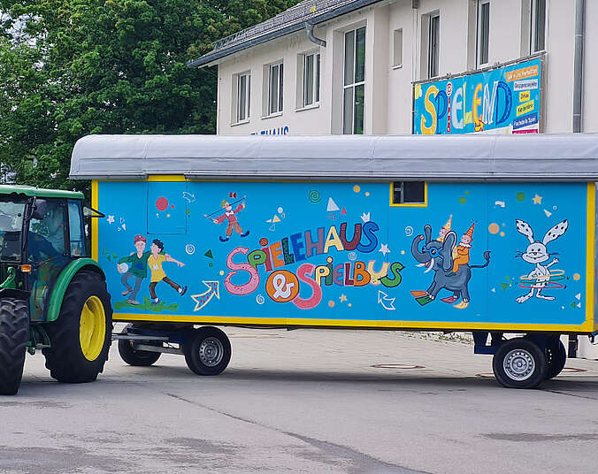 Bauwagen blau und mit bunten Bildern bemal hängt an einem grünen Traktor