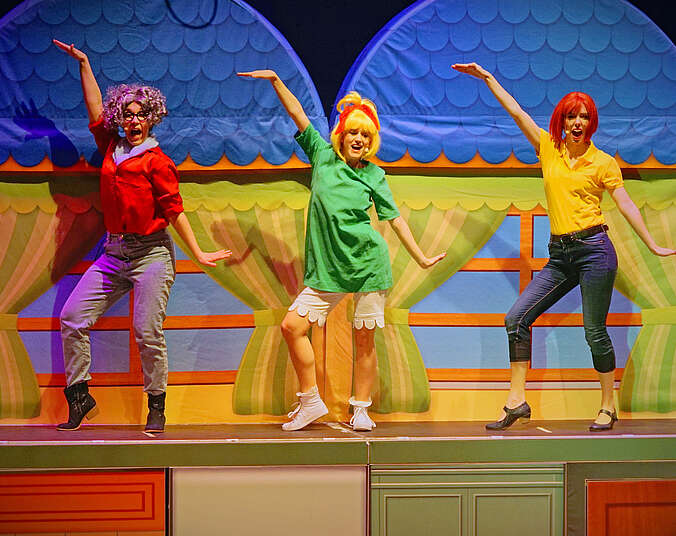 Drei Frauen tanzen und singen nebeneinander auf einem Podest. Im Hintergrund Ausschnitte aus Bühnenbild-Dach. Links Frau mit Locken und roter Bluse, Mitte blonde Bibi in grünem Kleid, rechts Mutter Barbara in 7/8-Jeans und gelber Bluse.