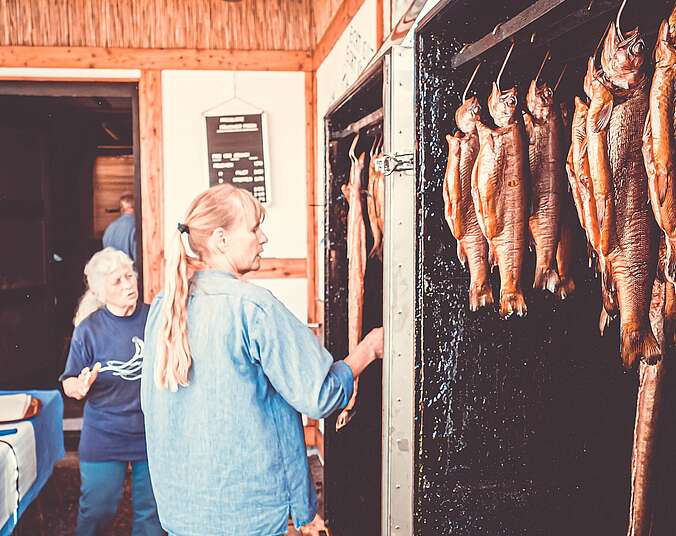 Eine Frau steht vor einem offenen Räucherofen. In diesem hängen geräucherte Fische.