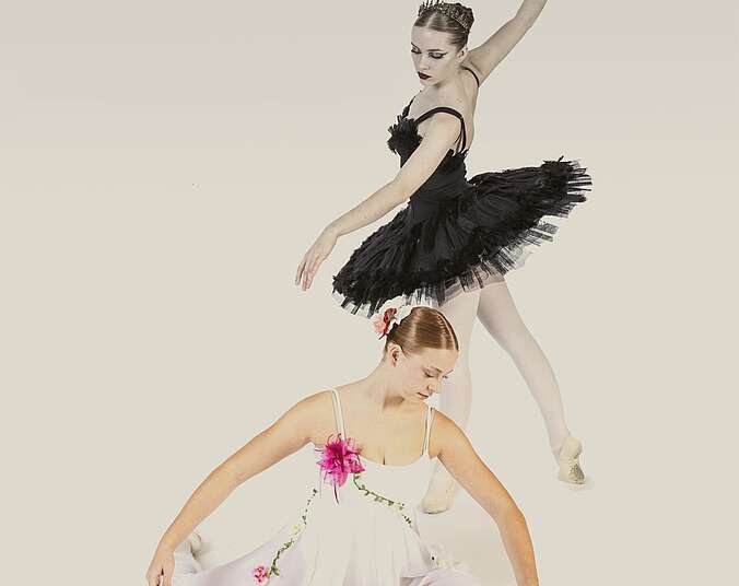 Zwei Baletttänzerinnen, eine stehend in schwarzem Tutu, die andere davor auf dem Boden in Ballettpose sitzend, in hellem Tanzkleid mit zwei pinken Blumen darauf.