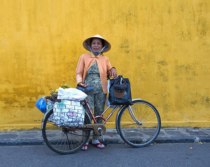 Eine lächelnde, ältere Frau asiatischer Herkunft in apricotfarbener Strickjacke, Stoffhose, Socken und roten FlipFlops mit spitzem Reishut steht hinter ihrem Fahrrad vor einer gelben Wand. Das Rad ist am Lenker und auf dem Gepäckträger beladen mit Plastiktüten und Gemüse.