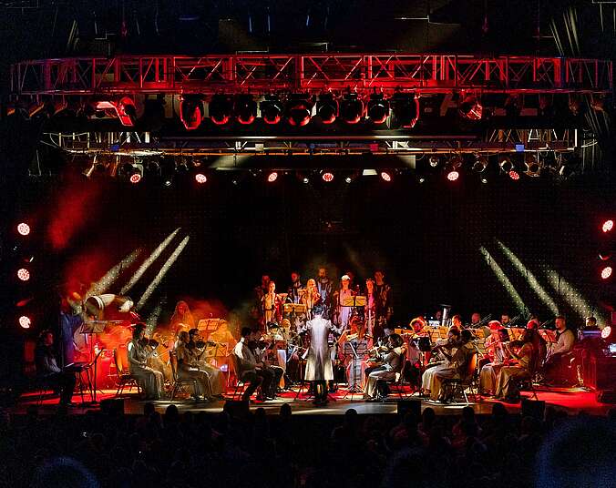 Ein Orchester mit Chor im Hintergrund auf einer rotbeleuchteten Bühne. Die Musiker werden von einem Dirigenten dirigiert, der einen langen silbernen Mantel trägt.