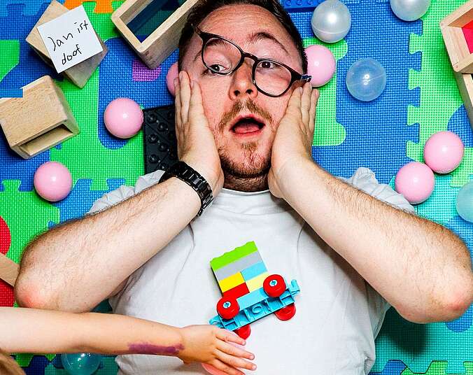 Ein Mann liegt auf einem bunten Spielteppich und ein Kind spielt mit Spielzeug auf ihm.