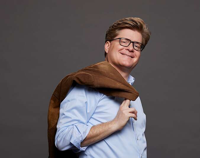 Ein Mann mittleren Alters mit braunen Haaren und Brille und freundlichem Lächeln blickt in die Kamera. Er trägt ein hellbblaues Hemd und hat eine braune Lederjacke über seine rechte Schulter geworfen welche er mit der Hand festhält.