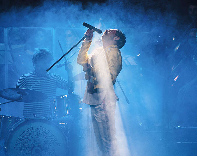 Ein Mann singt stehend in einen Mikrofonständer. Man erkennt nur seine komplette Silhouette im Profil; der Bühnenhintergrund ist blau und nebelig beleuchtet.