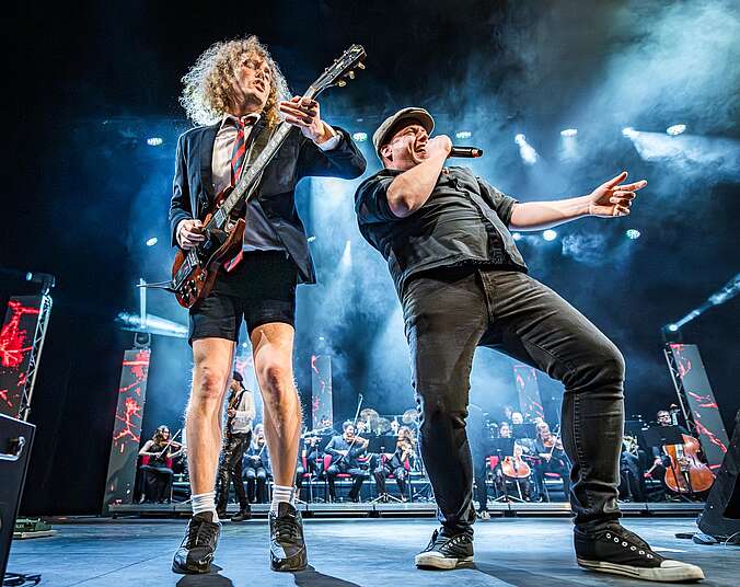 Zwei Männer stehen auf einer Bühne, einer hält eine Gitarre und der andere ein Mikrofon. Im Hintergrund erkennt man ein Orchester. Beide haben rockige Kleidung an und haben viel Spaß.