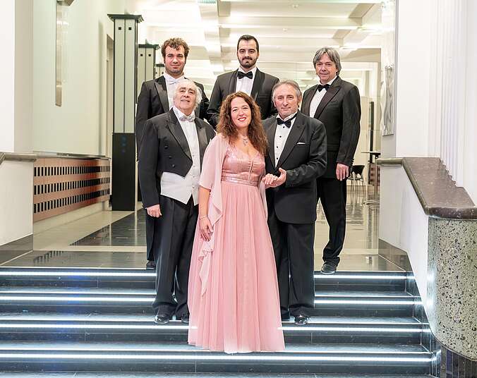 Fünf Männer gemischten Alters in schwarzen Galaanzügen stehen in einer Gruppe auf zwei unterschiedlichen Stufen einer Treppe. Auf der unteren Stufe in der MItte steht eine FRau mit brünetten Locken in rosa Abendkleid. Der Mann zu ihrer Linken hält galaant ihre Hand.
