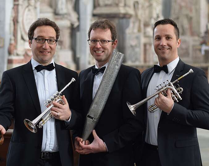 Drei Männer im schwarzen Anzug, halten Trompeten und eine Orgelpfeiffe hoch.