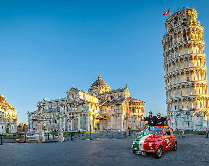 Ein alter FIAT 500 grün, weiß, rot gestreift mit der Aufschrift Luigui auf einem Platz vor alten italienischen Gebäuden, darunter ein schiefer Turm und zwei Männer in schwarzen Shirts.