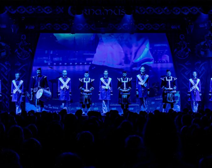 Sieben Tänzerinnen und Tänzer sowie vier Trommler jeweils in Uniformen mit weißen Kreuzen auf der Brust stehen in einer Reihe auf einer schwach in dunkelblauem Licht beleuchteten Bühne.
