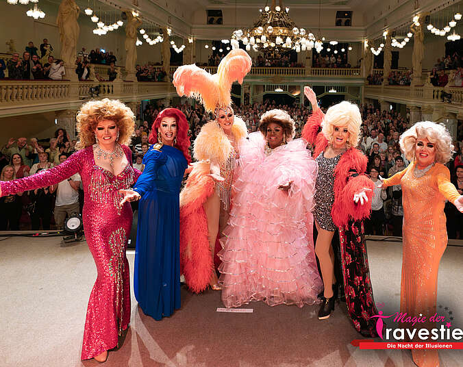 Sechs Travestie-Künstlerinnen stehen nebeneinander in bunten Paillettenkleidern auf einer Bühne in einem prunkvollen Saal sie drehen dem Saal den Rücken zu. Im Saal klatscht stehend begeistertes Publikum.