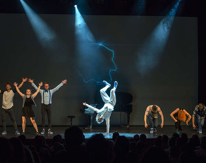 Auf einer Bühne stehen links drei Breakdancer, davon eine Frau, mit nach oben gestreckten Armen, in der Bühnenmitte macht ein männlicher Breakdaner einen einhändigen Handstand vor einem schwarzen Flügel, rechts auf der Bühne hocken drei Breakdancer, daruntre in der Mitte eine Frau in einer Reihe am Boden.