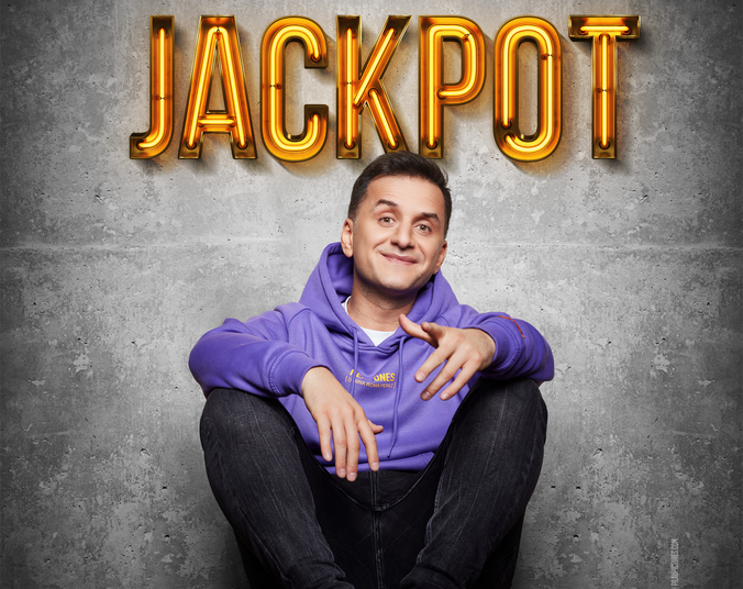 Ein Mann mit dichtem, dunklem Haar in lila Hoodie sitzt auf dem Boden vor einer Betonwand. Über ihm hängt in Neonbuchstaben das Wort "Jackpot".