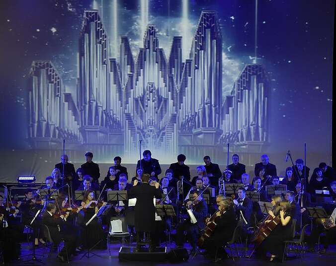 Ein in blau-violettes Licht getauchtes Orchester spielt auf einer Bühne. Im Hintergrund wird ein Bild einer stilisierter, sehr großen, silbrigen Orgel projeziert.