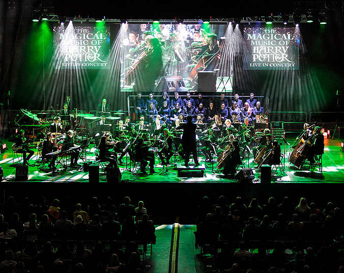 Ein Orchester spielt auf einer in grünem Licht beleuchteten Bühne. Im Hintergrund sitzt erhöht auf einem Podest ein Chor junger Menschen. Man kann die vorderen Reihen des Publikums von hinten erkennen.