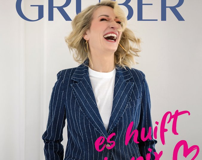 Eine blonde Frau mit welligem Haar mit offenem Mund herzlichen lachend. Sie trägt einen blauen Nadelstreifenanzug.