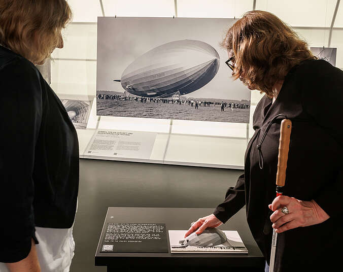 zwei Frauen in einer Ausstellung schauen sich ein Exponat an. Eine Frau ist blind und hält einen Langstock