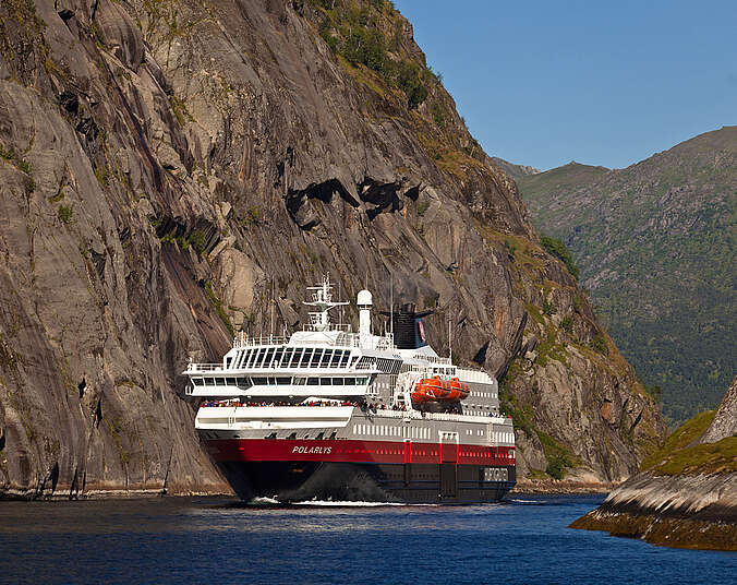 Ein Polar-Kreuzfahrtschiff mit blau-rotem Rumpf und weißem Aufbau färht durch einen Fjord, links und rechts gehen steile Felswände nach oben. In den oberen Etagen stehen vorne die Passagiere und schauen in die Landschaft.