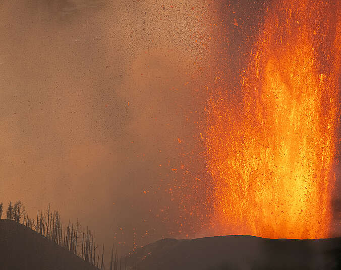 Orange-gelb leuchtende Lava spritzt fontänenartig aus einem schwarzen Hügel. Auf dem benachbarten Hügel stehen einige verbrannte Bäume, der Himmel ist nebelig-grau.