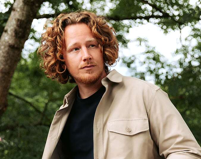 Ein junger Mann mit schulterlangen gelocken roten Haaren und Dreitagebart in schwarzem T-Shirt mit sandfarbener Jacke darüber; im Hintergrund Birkenbäume.