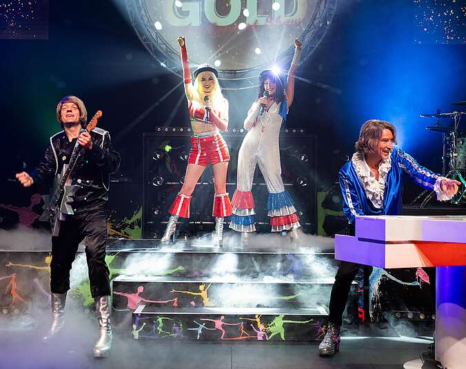 Zwei Männer an Guitarre und Keyboard links und rechts und zwei singende Frauen auf einer Showtreppe in der Mitte tragen schillernde Kostüme aus den 70er Jahren.