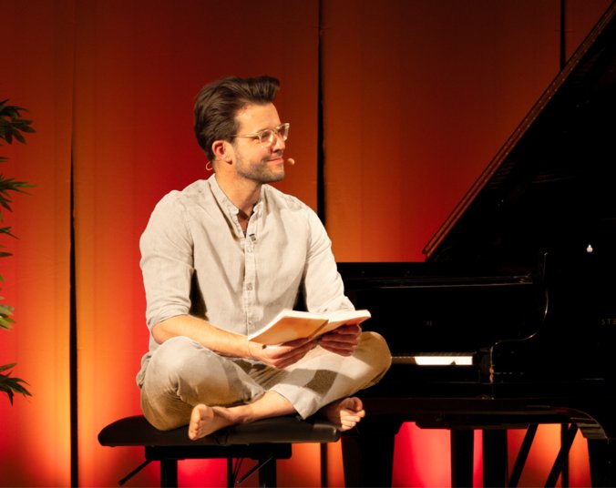 Ein junger Mann mit Brille in beigem Hemd und beiger Hose sitzt im Schneidersitz au einem Klavierhocker vor einem Flügel. Er hält ein aufgeschlagenes Buch in den Händen.