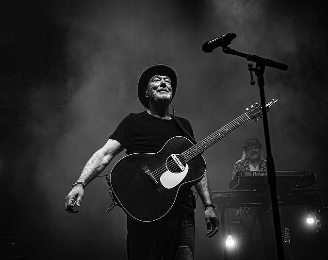 Älterer Mann in schwarzem T-Shirt, schwarzer Jeans und schwarzem Hut steht mit einer schwarzen Akustikgitarre vor einem Mikrofonständer auf einer Bühne. Im Hintergrund ist Nebel und ein ebenfalls älterer Mann mit grauem langen Locken und Hut hinter einem Keyboard zu sehen.