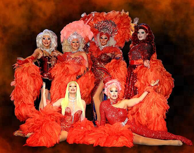 Sechs in orangenen Federkleidern Transvestiten strecken ihre Arme in die Luft und schauen dabei freundlich in die Kamera. Sie tragen verschiedene Perücken und sind alle stark geschminkt