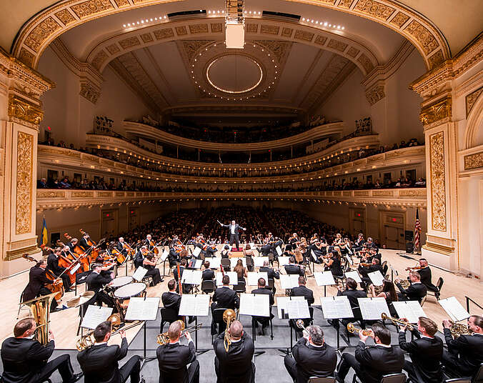 Ein Orchester auf der Bühne, fotografiert von der Bühne ins Publikum in den prunkvollen Saal der Carnegie Hall.