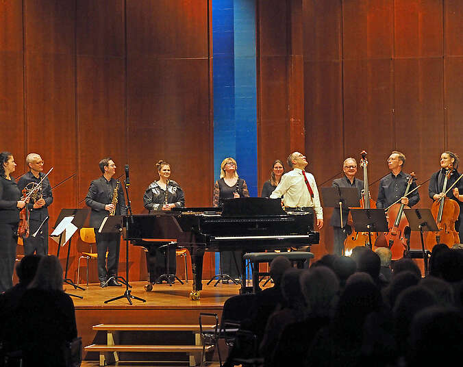 Orchester aus neun Musikerinnen und Musiker in dunkler Konzertkleidung mit ihren jeweiligen Instrumenten in der Hand stehen auf einer Bühne um einen Mann in weißem Hemd mit roter Krawatte. Der Mann steht an einem Flügel.