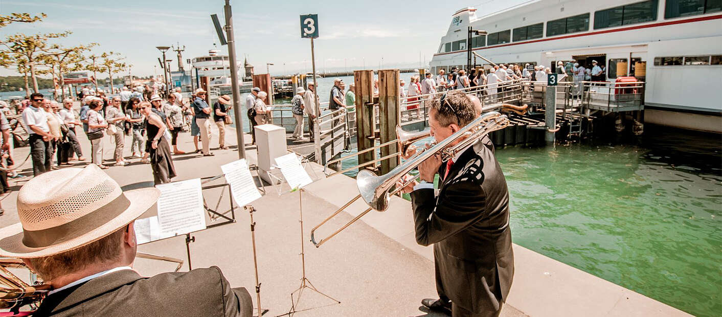Bläsertrio steht an Ufer vor Fähre und spielt, während Menschen anstehen um auf die Fähre zu gelangen.