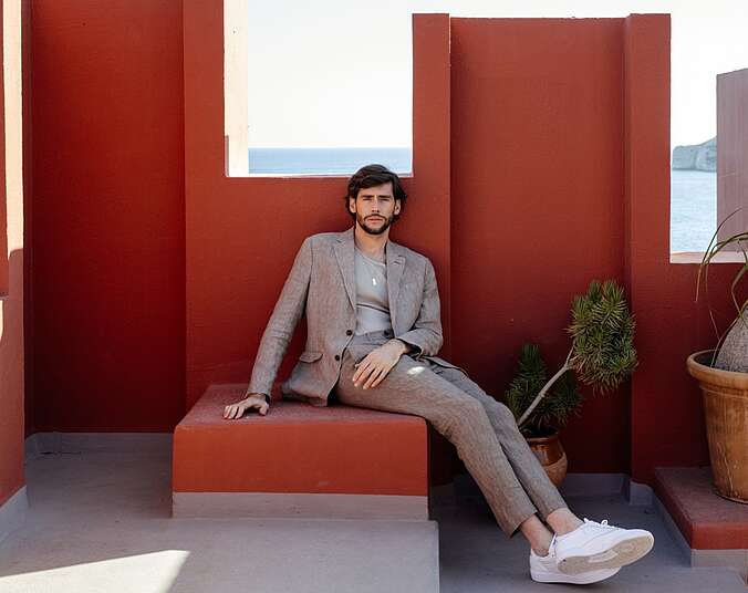 Ein junger Mann mit braunen Haaren trägt einen grauen, modernen Anzug mit weißen Turnschuhen. Er sitzt entspannt auf einer Stufe und lehnt sich an eine terrakottafarbene Wand. Durch die Fensteröffnungen des mediterranen Gebäudes sieht man das Meer.