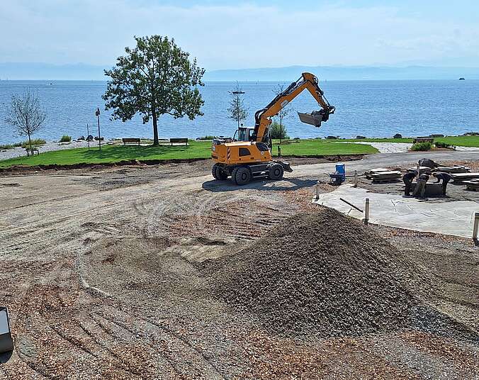Ein gelber Bagger auf einer Schotterfläche neben einer mit Steinplatten gelegten Außenterrasse. Im Hintergrund liegt der Bodensee bei schönem Wetter.