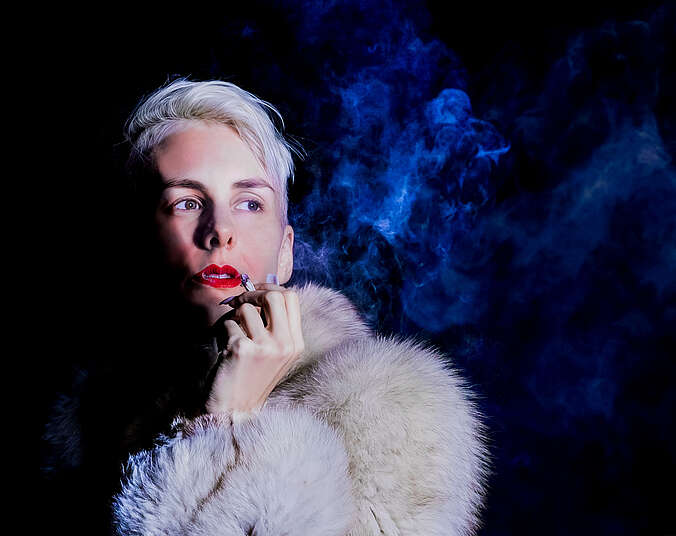 Eine Frau mit hellblonden, kurzen Haaren hält eine Zigarette in der Hand und schaut ins Leere. Dabei trägt sie einen hellgrauen Pelzmantel und roten Lippenstift