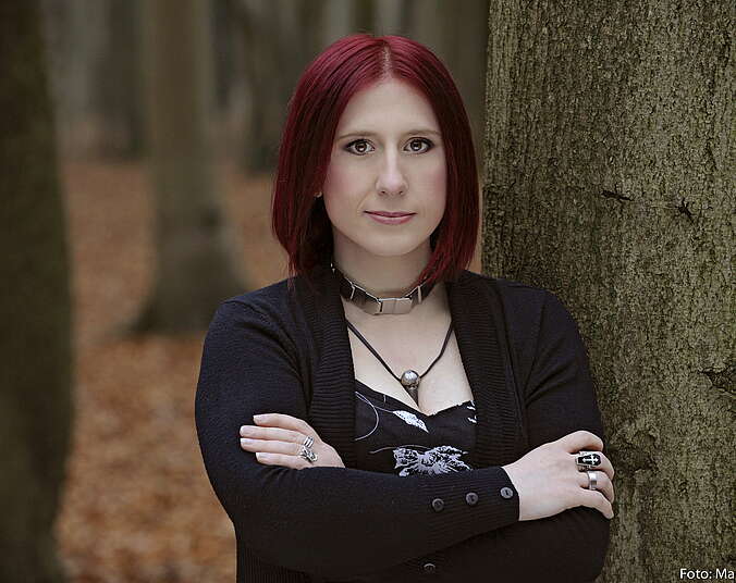 Eine junge Frau, schwarz gekleidet, mit rot gefärbten Haaren, Silberringen und Silberhalsband steht mit verschränkten Armen an einem Baum im Wald. Sie schaut den Betrachter freundlich an.