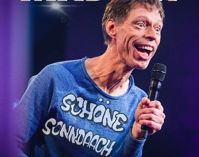 Ein Mann mittleren Alters mit sehr großem Mund trägt einen blauen Pullover mit der Aufschrift "Schöne Sonndaach". Er hält ein Mikrofon in seiner linken Hand.
